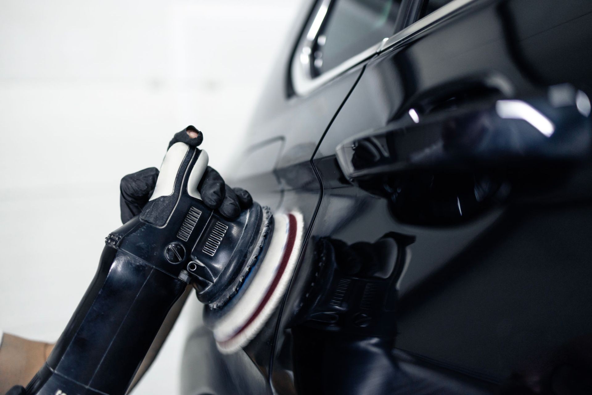 Person using a machine polisher on a shiny black car door, wearing black gloves.
